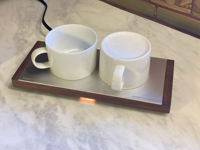Two white ceramic mugs resting on a temper cup pre-heater with a dark American walnut frame and aluminum plate, sitting on a white marble countertop.