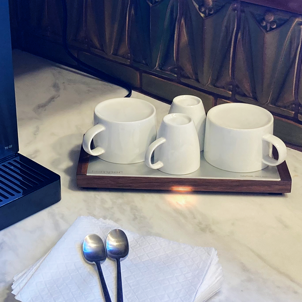 A set of four white ceramic coffee mugs on a heated tray, placed on a marble countertop next to two metal spoons on a napkin.