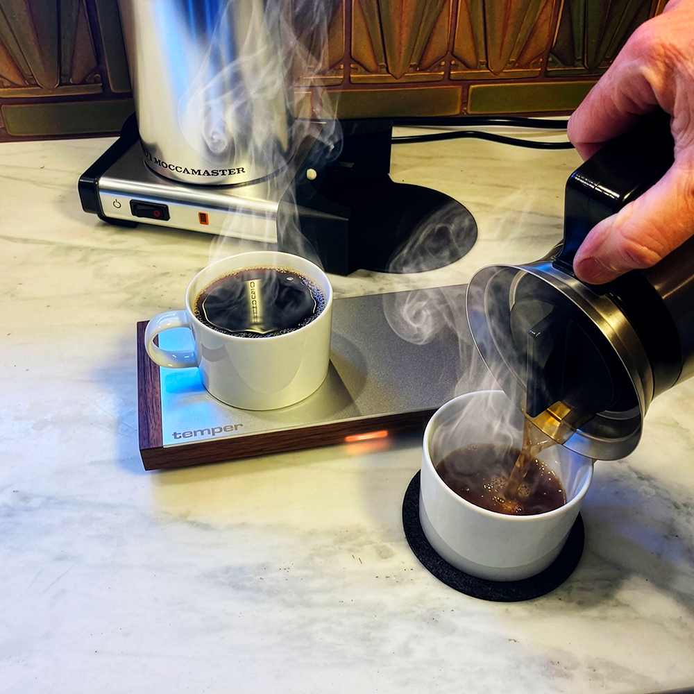 A hand pours freshly brewed coffee from a black and stainless steel pour-over maker into a white cup on a marble countertop. Another white cup sits on a temper cup pre-heater, and a coffee maker is visible in the background.