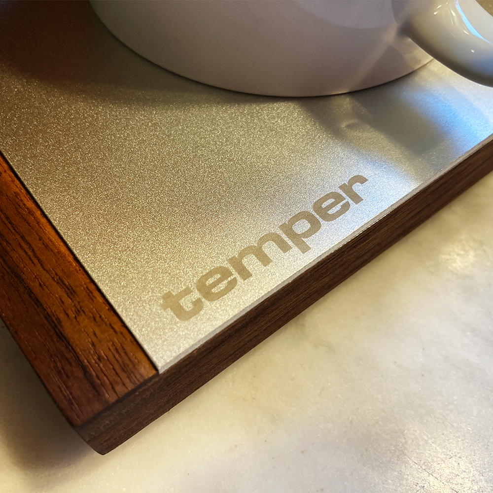 Close-up of a temper cup pre-warmer with the brand name 'temper' laser etched on the surface, with a white ceramic mug partially visible at the top.